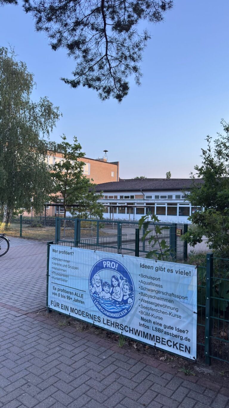 Vereinsbanner „Für ein modernes Lehrschwimmbecken“ am Zaun der Lerchenschule Faßberg, dahinter das Schulgebäude mit der Schwimmhalle