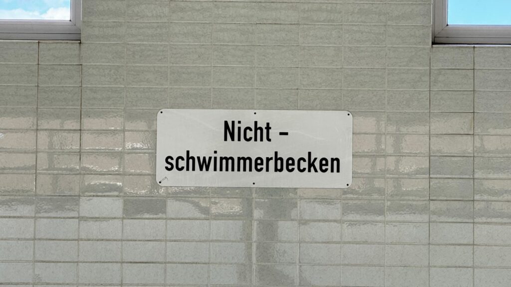 Schild mit der Aufschrift „Nichtschwimmerbecken“ an der gefliesten Wand der Schwimmhalle