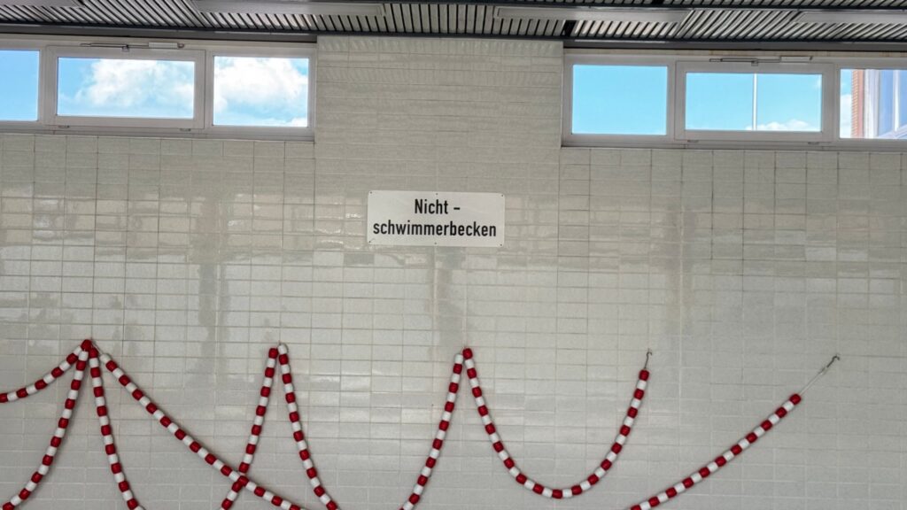 Geflieste Hallenwand mit Oberlichtern, dem Schild „Nichtschwimmerbecken“ und aufgehängten rot-weißen Schwimmleinen