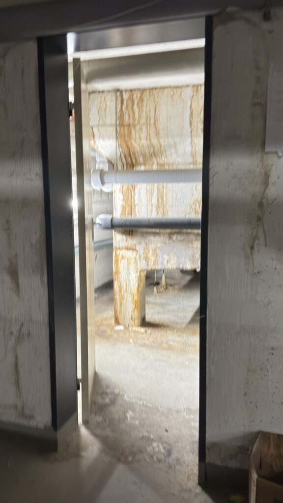 Blick durch eine Tür in einen Technikgang mit Ablaufspuren an den Betonwänden